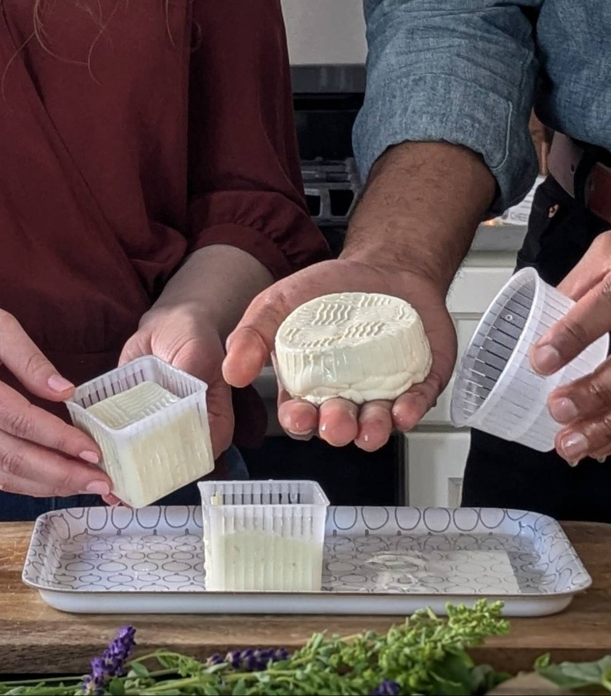 Two people holding small containers of cheese with a loaf of bread and herbs on a table.