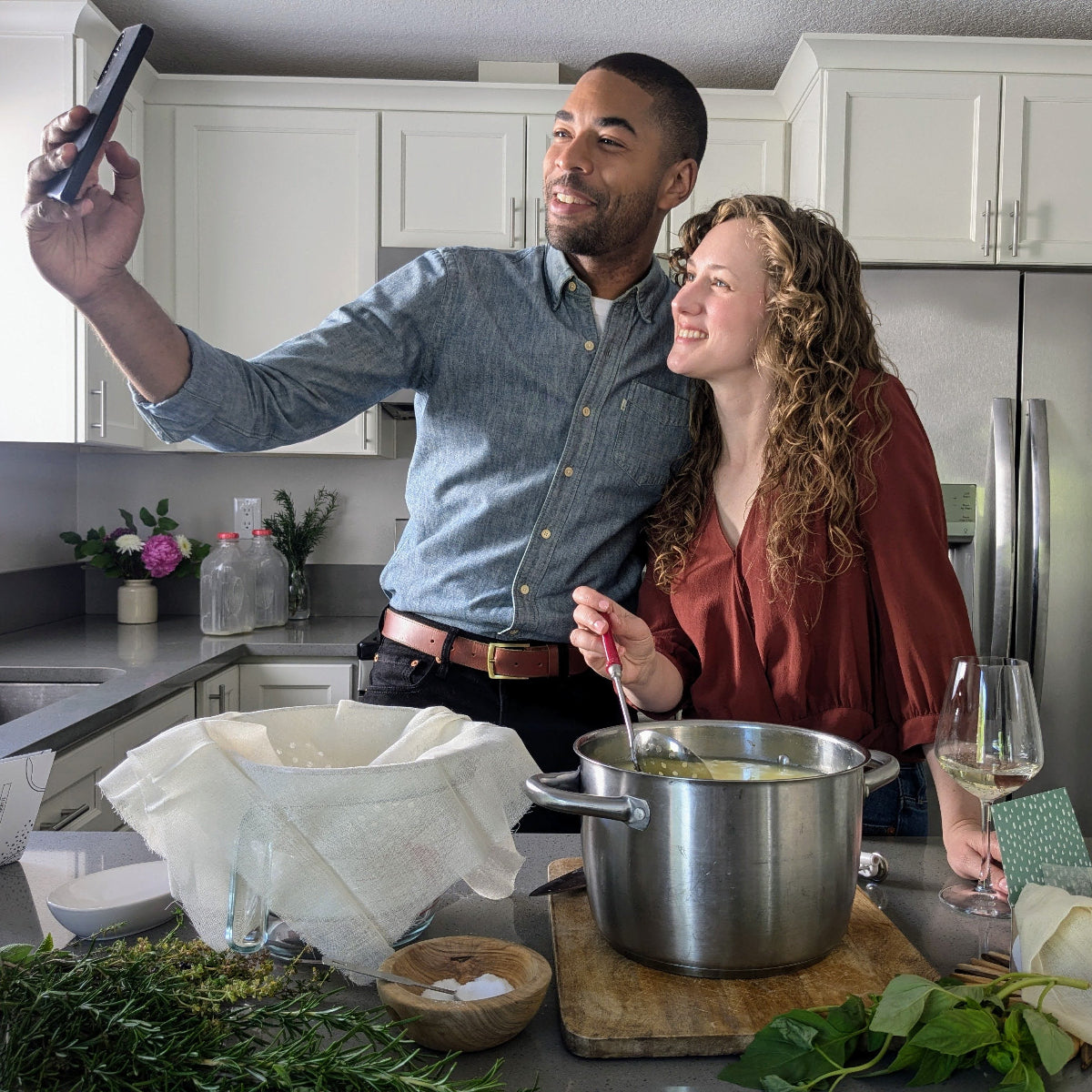 young couple in a kitchen making cheese. The man is holding a phone up to take a selfie of themselves and their date night activity.