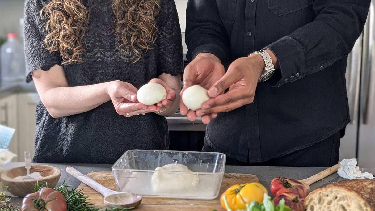 Two people holding their homemade mozzarella in a kitchen with ingredients and appliances around