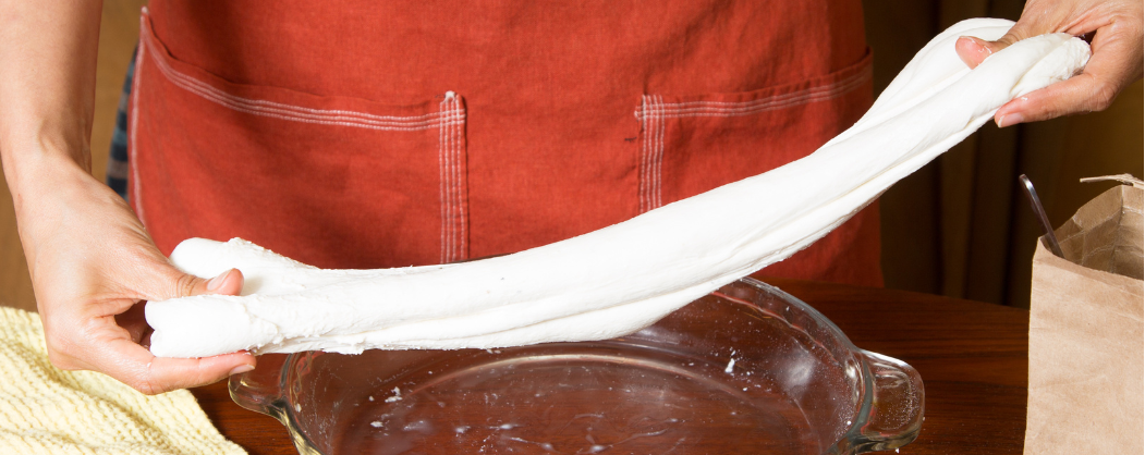 Person in a red apron stretching white dough over a glass pie dish on a wooden table.