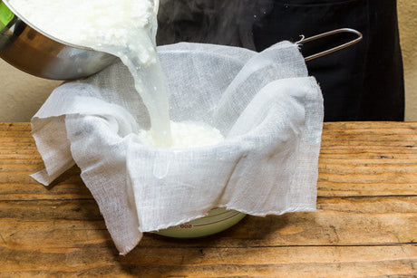 cheesecloth draped over a colander and bowl on a wood counter with curds and whey being pour in by someone in a black apron