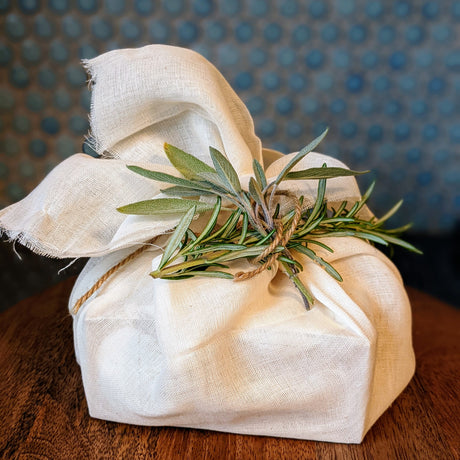 a cheesecloth wrapped mozzarella kit tied with natural twice and a spring of fresh green rosemary on wooden cheese tray in front of blue tile