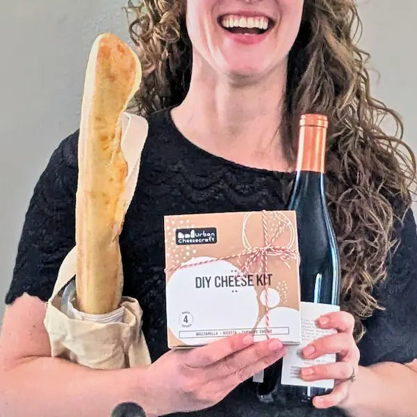 Person holding a bottle of wine, a baguette, and a DIY cheese kit against a plain background