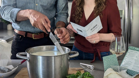 couple cutting curd to make cheese. woman is reading instructions. man is holding a knife in a pot of milk. They are in a light colored kitchen. There is a cheese kit box, herbs and a cutting board nearby.