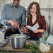 couple cutting curd to make cheese. woman is reading instructions. man is holding a knife in a pot of milk. They are in a light colored kitchen. There is a cheese kit box, herbs and a cutting board nearby.
