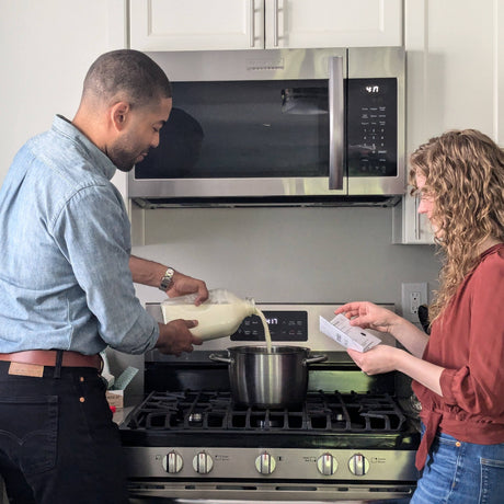 couple at a stove pouring milk into a stainless steel pot. Man is holding the glass bottle of milk over the pot, woman is reading cheese making instructions.