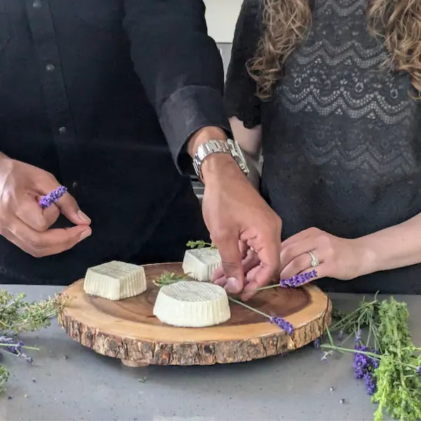 Two people arranging cheese on a wooden board with lavender flowers.